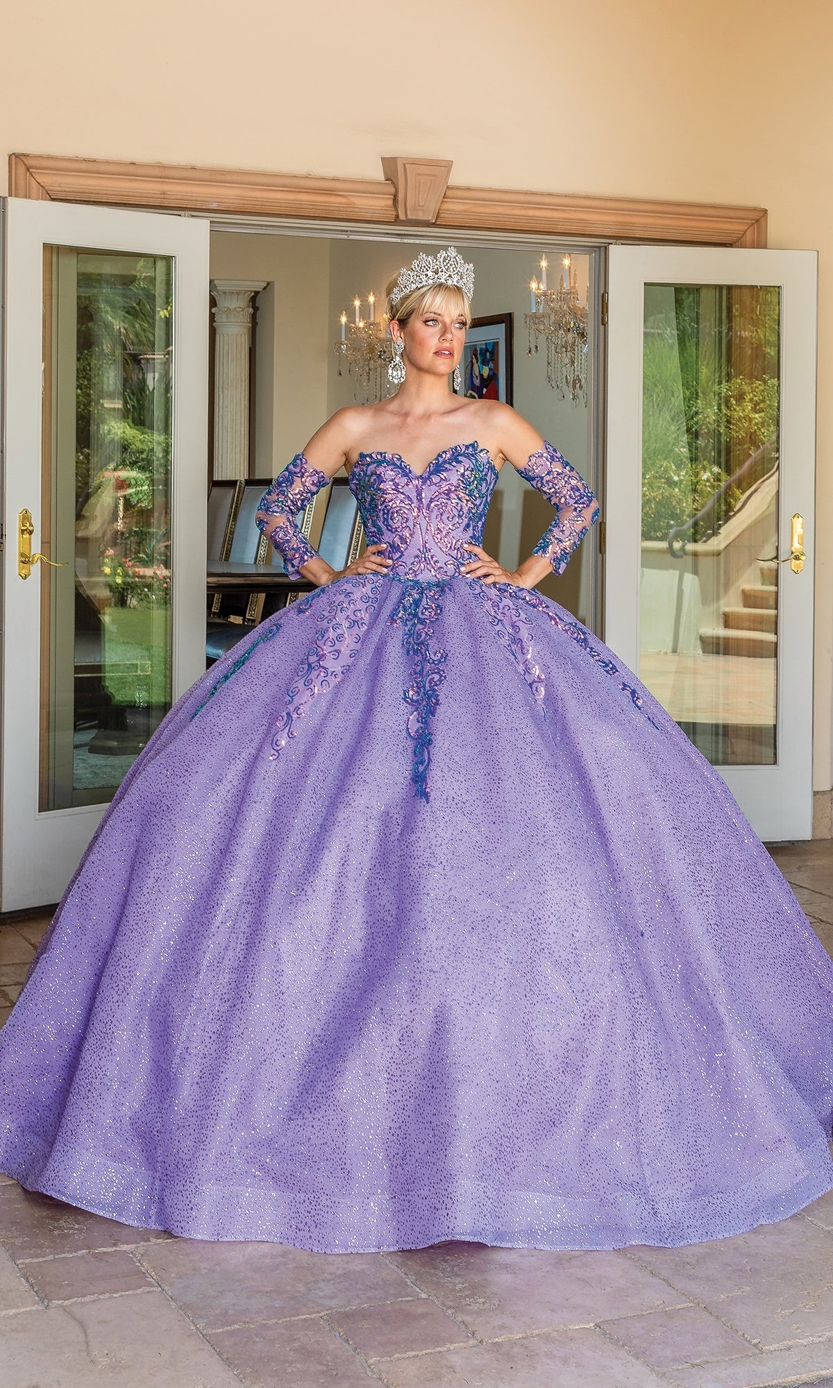 Wearing the Quinceanera Dress 1675 By Dancing Queen, a sparkling lavender ball gown with blue and pink embroidery, matching gloves, and a tiara, she stands in front of open glass doors.